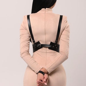 May include: A woman wearing a nude-colored dress with a black leather harness and matching wrist cuffs. The harness has a buckle and metal accents.
