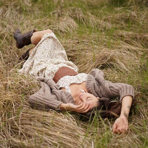 May include: A person wearing a floral print dress, brown corset, and brown boots, lying in a field of tall grass. A brown knit cardigan is draped over the person's arms. The scene is set outdoors in natural light.