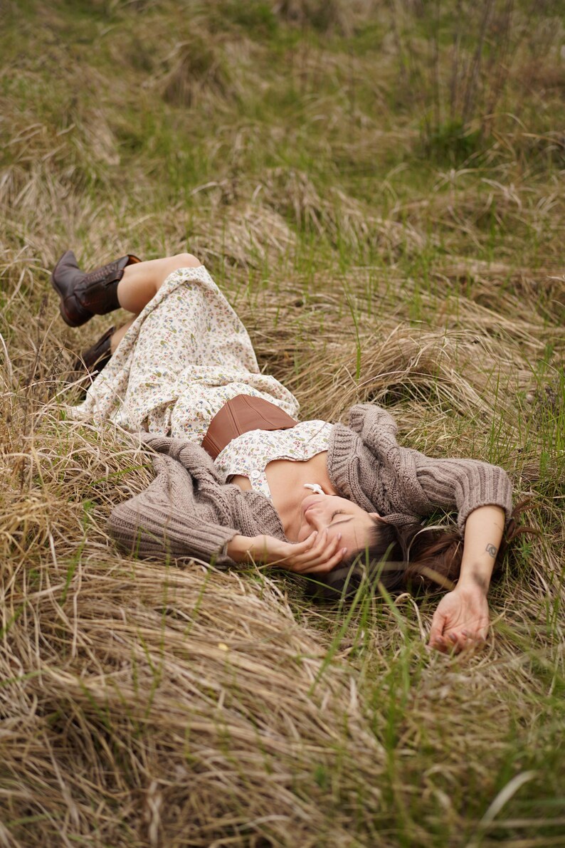 May include: A person wearing a floral print dress, brown corset, and brown boots, lying in a field of tall grass. A brown knit cardigan is draped over the person's arms. The scene is set outdoors in natural light.