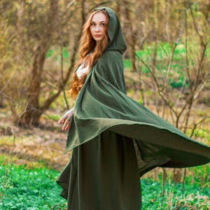 Può includere: Un lungo mantello verde foresta con cappuccio. Il mantello è indossato all'aperto in una zona boscosa con erba verde e alberi. Il mantello è realizzato in un tessuto morbido ed è adatto per eventi di cosplay o in costume.