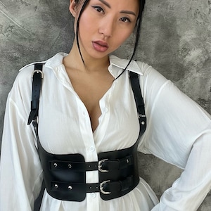 May include: A black leather harness with silver buckles and straps. The harness is worn over a white button-down shirt.