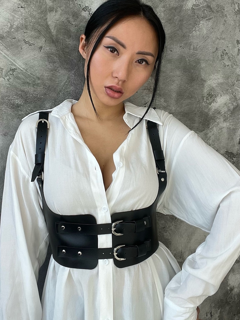 May include: A black leather harness with silver buckles and straps. The harness is worn over a white button-down shirt.