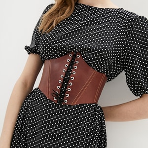 May include: A brown leather corset with a lace-up front. The corset is worn over a black polka dot dress.