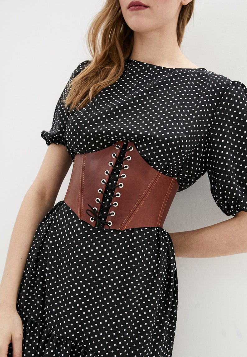 May include: A brown leather corset with a lace-up front. The corset is worn over a black polka dot dress.