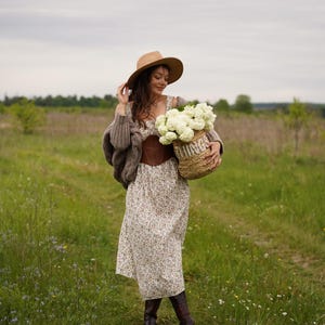 May include: A woman in a field wearing a floral dress, brown corset, and hat, holding a woven basket filled with white flowers. She is also wearing a brown sweater and boots. The basket has the word "THINGS" on it.