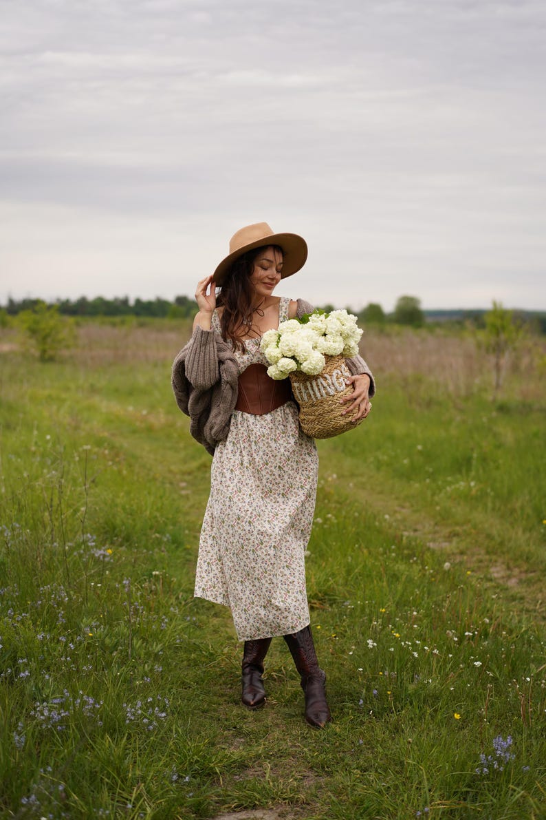 May include: A woman in a field wearing a floral dress, brown corset, and hat, holding a woven basket filled with white flowers. She is also wearing a brown sweater and boots. The basket has the word "THINGS" on it.