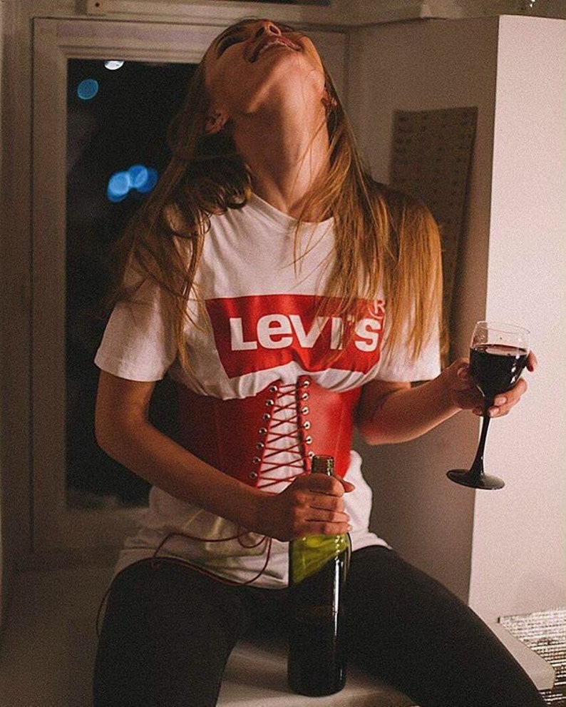 May include: A woman wearing a white t-shirt with the word "Levi's" printed on it, a red leather corset, and black pants. She is holding a bottle of wine and a glass of red wine.
