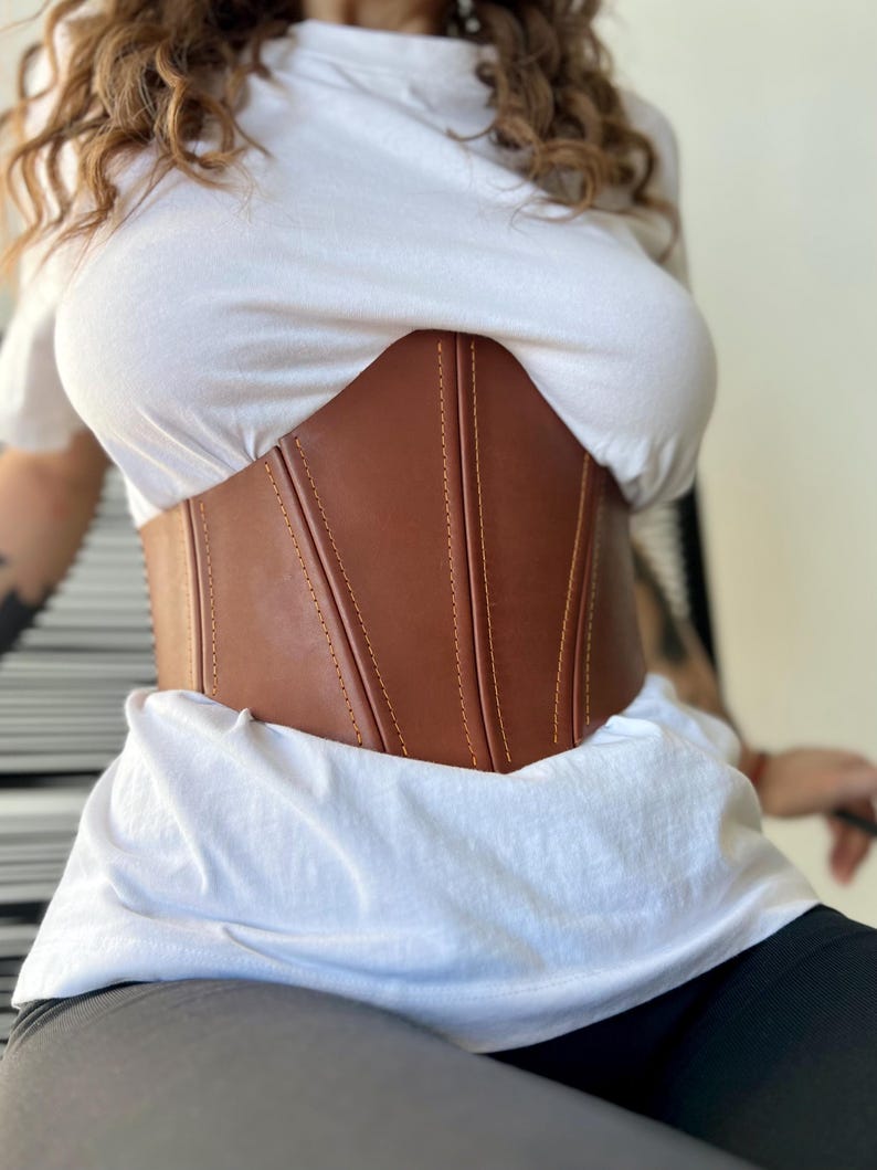 May include: A brown corset with vertical seams and yellow stitching. The corset is worn over a white t-shirt. The corset is made of a smooth material and has a structured design.