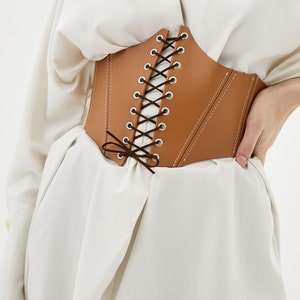 May include: A brown leather corset with a lace-up front and a bow tie at the bottom. The corset has a white stitching detail.