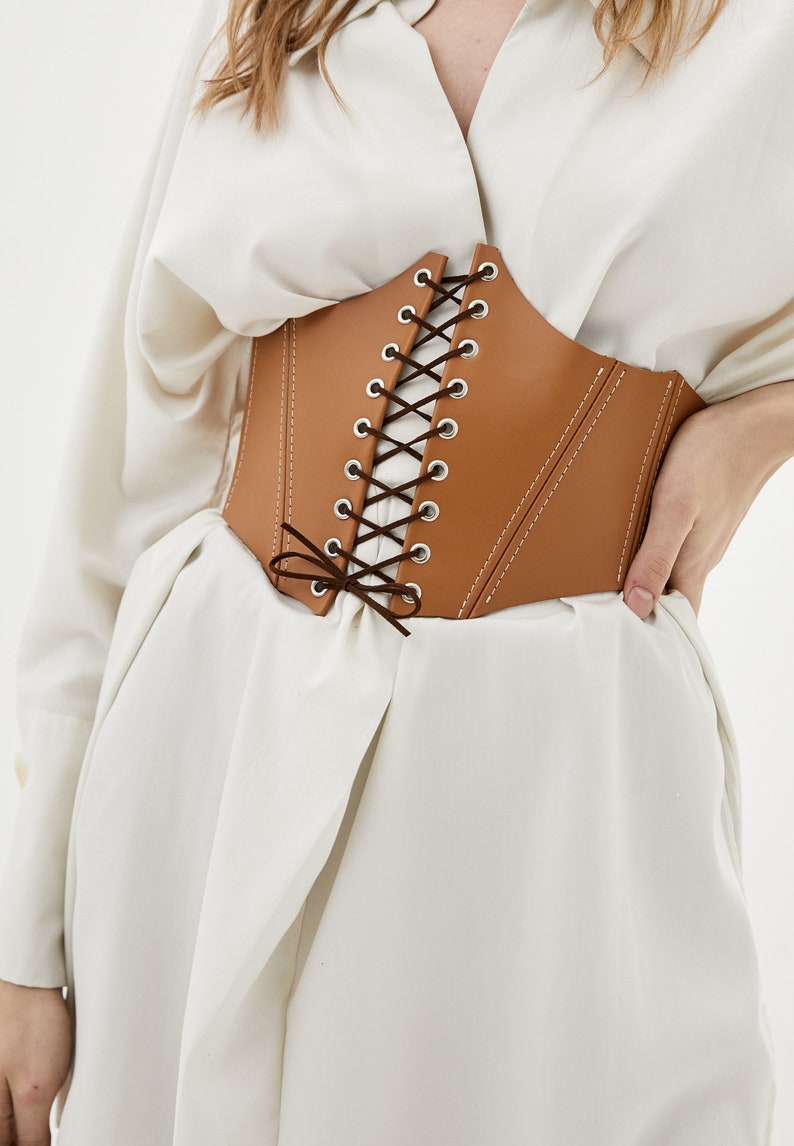 May include: A brown leather corset with a lace-up front and a bow tie at the bottom. The corset has a white stitching detail.