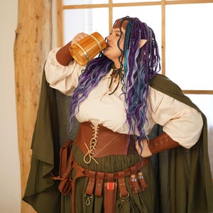 May include: A person in a fantasy-themed outfit drinks from a wooden mug. The ensemble includes a cream-colored blouse, brown corset, and a green skirt and cape. Accessories include a brown belt with pouches and small bottles. The person has purple and blue hair.