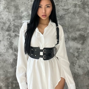 May include: A black leather harness with double buckles and silver accents. The harness is worn over a white button-down shirt.