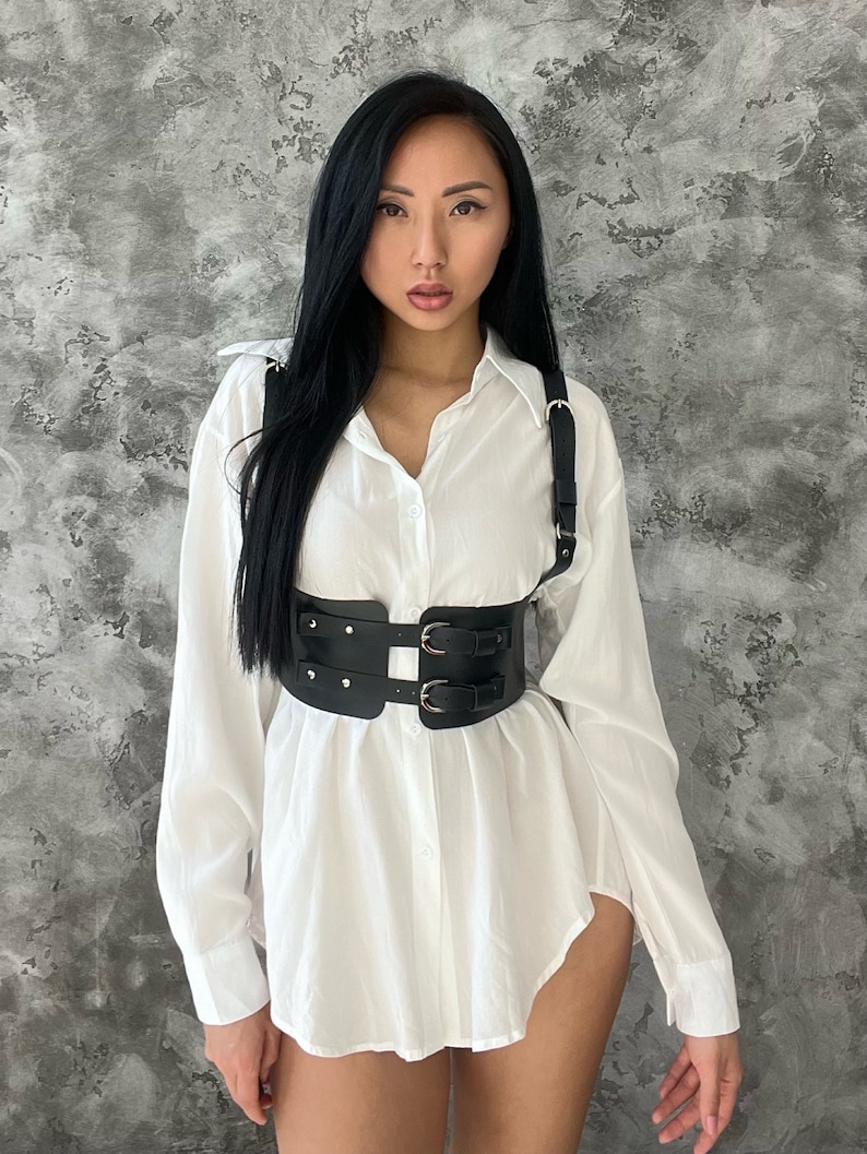 May include: A black leather harness with double buckles and silver accents. The harness is worn over a white button-down shirt.