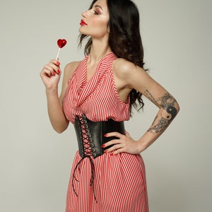 May include: A woman wearing a red and white striped dress with a black leather corset belt. She is holding a red heart-shaped lollipop and has a tattoo on her arm.