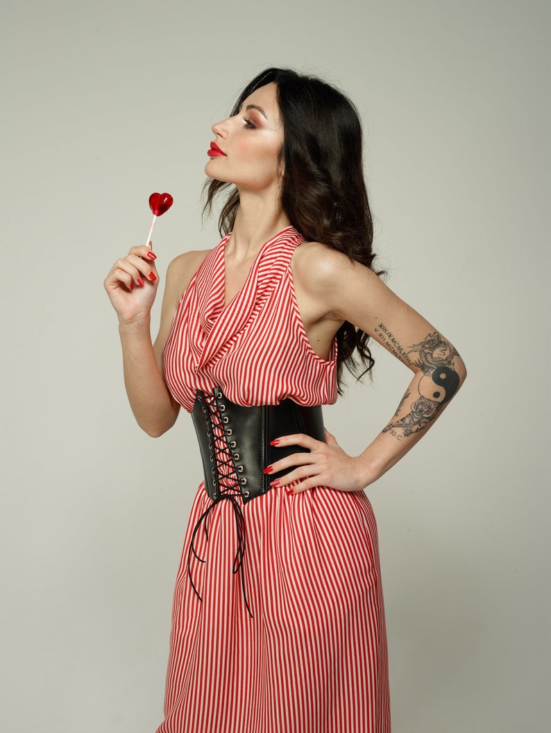 May include: A woman wearing a red and white striped dress with a black leather corset belt. She is holding a red heart-shaped lollipop and has a tattoo on her arm.