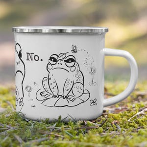 May include: White enamel mug with a black and white illustration of a grumpy frog on a lily pad, with the word "No." and a fly. The mug has a silver rim and a white handle. The background is blurred.
