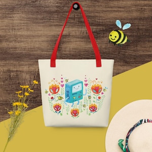 Puede incluir: Bolso tote crema con un diseño floral colorido y un personaje de dibujos animados. El bolso tiene asas rojas y cuelga sobre un fondo de madera. También se ve una abeja amarilla y negra.