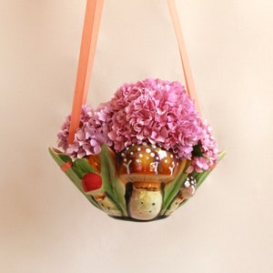 Op de afbeelding: Een roze en groene keramische hangende plantenpot in de vorm van paddenstoelen met roze hortensia bloemen die hangen aan perzikkleurige linten.