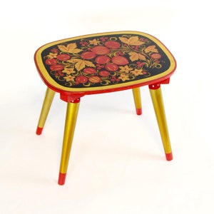 Puede incluir: Una pequeña mesa de madera pintada de rojo y dorado con una tapa negra con un diseño floral. La mesa tiene cuatro patas.