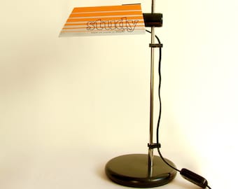 May include: A retro study lamp in black and chrome, featuring an orange and silver shade with the word "study". The lamp has a circular base and a long, adjustable arm. The lamp is plugged into a wall outlet.