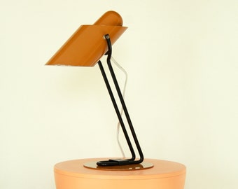 May include: A mid-century modern desk lamp with a black metal base and an orange metal shade. The lamp is sitting on a peach-coloured table.