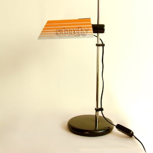 Könnte beinhalten: Vintage Schreibtischlampe in Schwarz und Chrom mit orangefarbenem und silberfarbenem Schirm. Auf dem Schirm steht das Wort "study". Die Lampe hat einen runden Fuß und einen langen, verstellbaren Arm. Die Lampe ist eingesteckt.