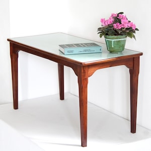 May include: A rectangular wooden table with four tapered legs and a light-colored top surface. A book titled Paradiso by Antonio Perazzi and a green ceramic pot containing a blooming pink African violet plant are placed on the table surface.