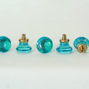 Puede incluir: Siete pomos de armario de vidrio azul turquesa con bases de metal dorado. Los pomos tienen una forma ligeramente redondeada y abovedada.
