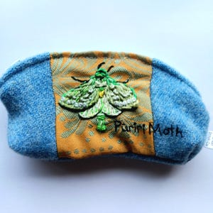 May include: A zipped pouch featuring blue textured fabric on the sides and a central orange panel with a green embroidered moth. The text Puriri Moth is stitched in black thread below the moth, and a small white tag reads Blanket Stitch Co.