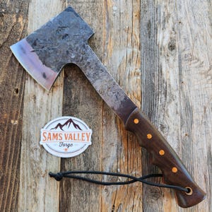 May include: A handcrafted axe with a dark wooden handle and a metal head. The axe head has a unique shape and a worn appearance. The handle is adorned with brass rivets. The axe is displayed on a wooden surface, with a sticker that reads "Sams Valley Forge".