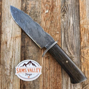 May include: A large, handcrafted knife with a textured, wavy blade and a dark wooden handle. The blade has a unique pattern, and the handle is secured with a metal bolster. A sticker with the text "Sams Valley Forge" is also visible.