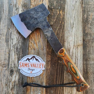 May include: A handcrafted hatchet with a dark, textured blade and a handle featuring a colorful wood grain pattern in shades of orange, green, and brown. The hatchet rests on a wooden surface, with a sticker that reads "Sams Valley Forge".