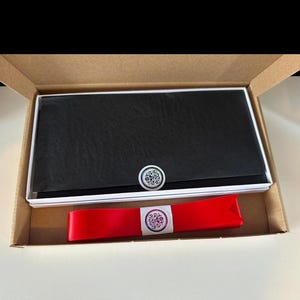 K&ouml;nnte beinhalten: Eine schwarze Geldb&ouml;rse mit dekorativem Knopf wird in einer Pappschachtel pr&auml;sentiert. Ein rotes Band mit dekorativem Etikett ist ebenfalls enthalten. Die Geldb&ouml;rse ist rechteckig und scheint aus Leder zu sein.