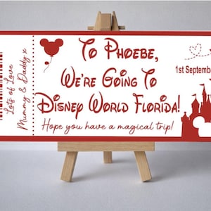 Op de afbeelding: Een rood en wit ticket dat een reis naar Disney World Florida aankondigt. De tekst luidt "To Phoebe, We're Going To Disney World Florida!" met een Mickey Mouse-silhouet en de datum "1 september 2025".