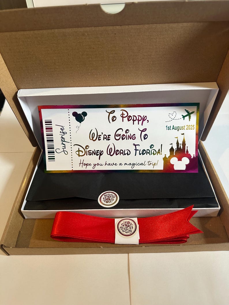 Op de afbeelding: Een cadeauverpakking met een Disney World Florida-ticket. Op het ticket staat "To Poppy, We're Going To Disney World Florida!" met de datum "1 augustus 2025". Een rood lint en een decoratieve sticker zijn ook inbegrepen.