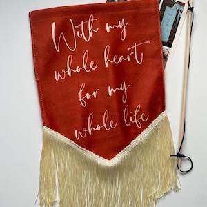 Peut inclure: Une bannière de couleur rouille avec l'inscription "With my whole heart for my whole life" en écriture blanche. La bannière est ornée d'une frange crème et d'une baguette en bois. Un magazine intitulé "The Banner Edit" est visible.
