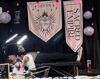 May include: A tattoo studio scene with an artist at work. Banners reading "TATTOOS BY TINA" and "SACRED EMPIRE" are displayed. The studio is decorated with paper lanterns and fairy lights. A sign displays the text "KUPS COME DAY RT 10 S 5 TS 10".