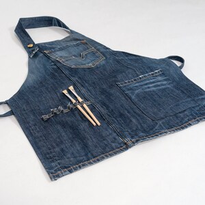 May include: A blue denim apron with a large pocket and two paintbrushes tucked into a smaller pocket.