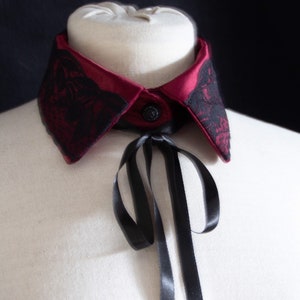 Könnte beinhalten: Ein burgunderfarbener Satin-Kragen mit schwarzer Spitzenborte und einer schwarzen Satin-Schleife. Der Kragen befindet sich auf einer weißen Puppe.