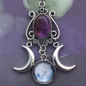 May include: A silver necklace with a pendant featuring a purple amethyst stone and three crescent moons. The pendant is ornate and detailed, with a delicate chain.