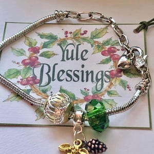 May include: A silver charm bracelet with multiple charms. Charms include a gold holly leaf, a green faceted bead, and a silver bead. The background features a card with the words "Yule Blessings" surrounded by a wreath of holly and berries.