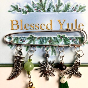 May include: A silver safety pin with dangling charms. Charms include a horn, a green bead, a starburst, black beads with a white bead and green leaf, and a holly leaf. The text "Blessed Yule" is visible.