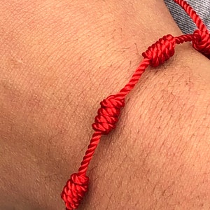Op de afbeelding: Een rood armband met geknoopte accenten. Het armband wordt om de pols gedragen.