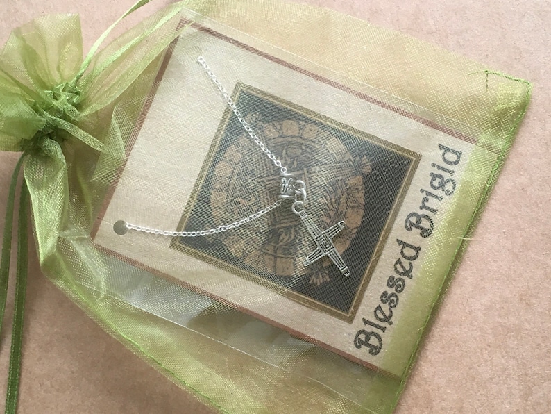 May include: A silver necklace with a Celtic cross pendant on a green organza bag with a card that reads "Blessed Brigid".