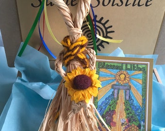 May include: A straw doll with a sunflower and colorful flowers on its head. The doll is in a box with blue tissue paper and a card that says "Blessed Litha".