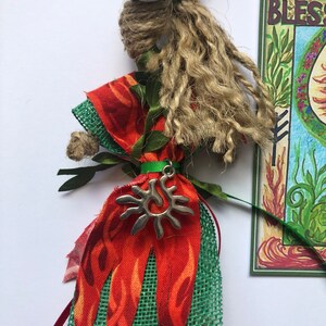 May include: A handmade doll with a long, brown, braided hair, wearing a red and green dress with a sun charm. The doll is standing in front of a card that says "Bless".