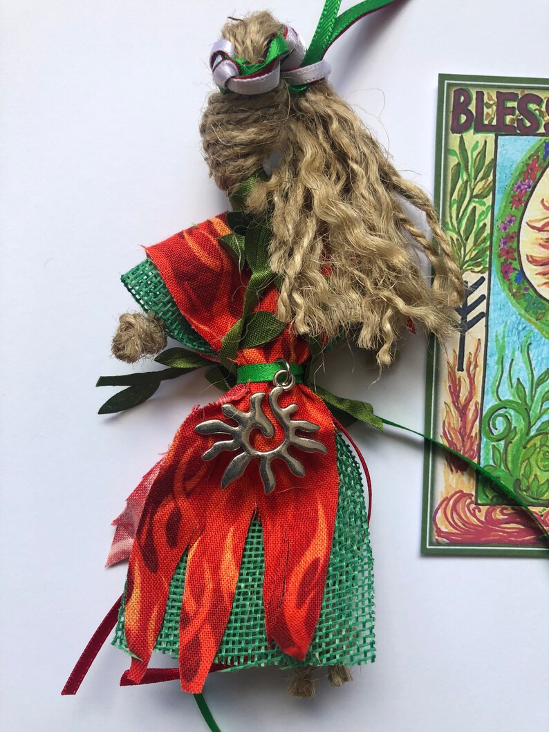 May include: A handmade doll with a long, brown, braided hair, wearing a red and green dress with a sun charm. The doll is standing in front of a card that says "Bless".