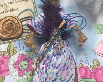 May include: A handmade doll with a jute body, wearing a patterned dress with purple accents and a faux fur collar. The doll has a braided staff and a silver charm. The background includes a card with text and floral illustrations.