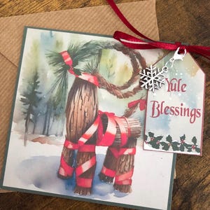 May include: A watercolor Christmas card featuring a Yule goat made of branches and tied with red ribbon. The card has a green border and a tag attached with the text "Yule Blessings" and a snowflake charm.