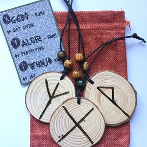 May include: Three wooden rune pendants with black cords and wooden beads. The runes are Gebō, Algiz, and Wunjo. The runes are on a red hessian bag with the meaning of each rune written on a card.